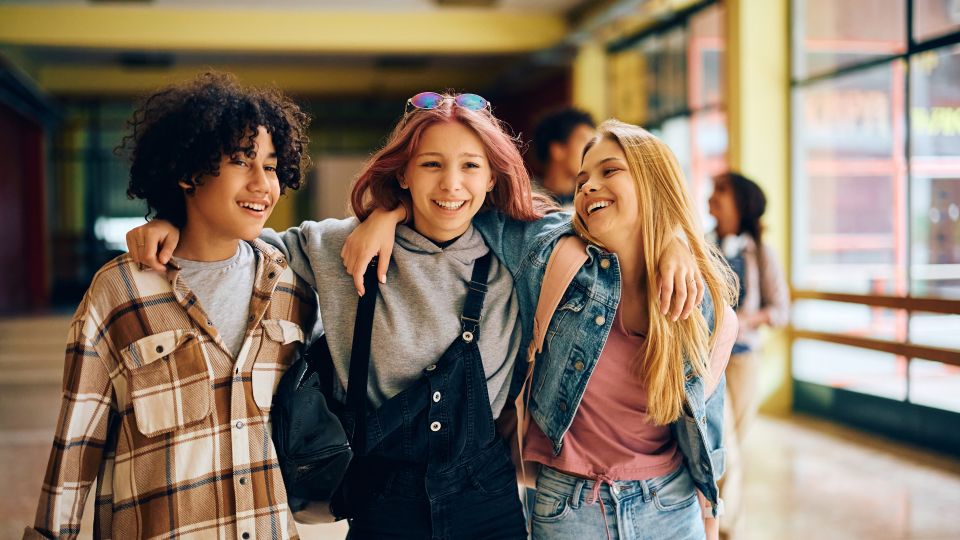 Three young adolescent friends with their arms around each other walking through a school hallway.